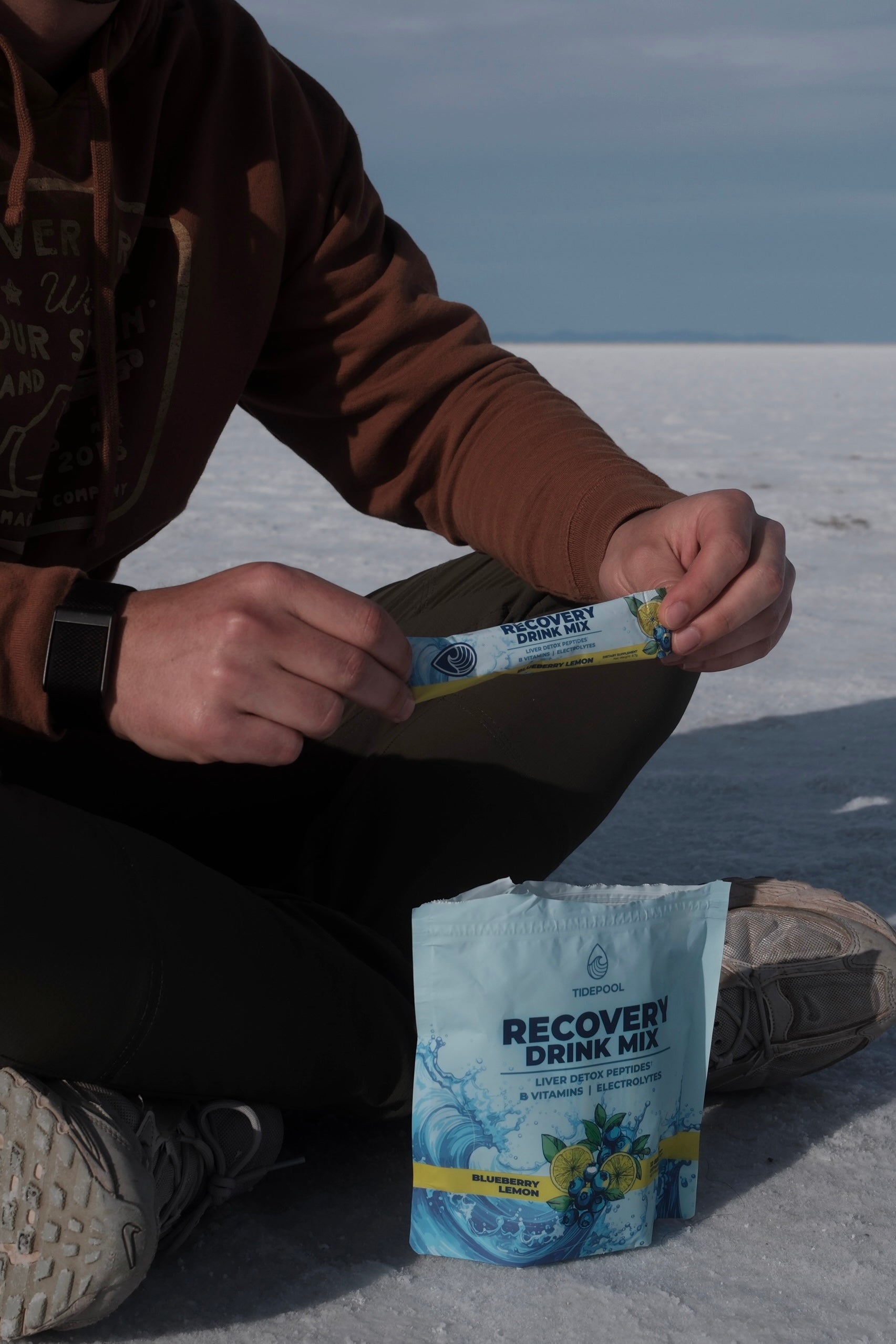 Person sitting on a snowy ground holding a recovery drink mix package.