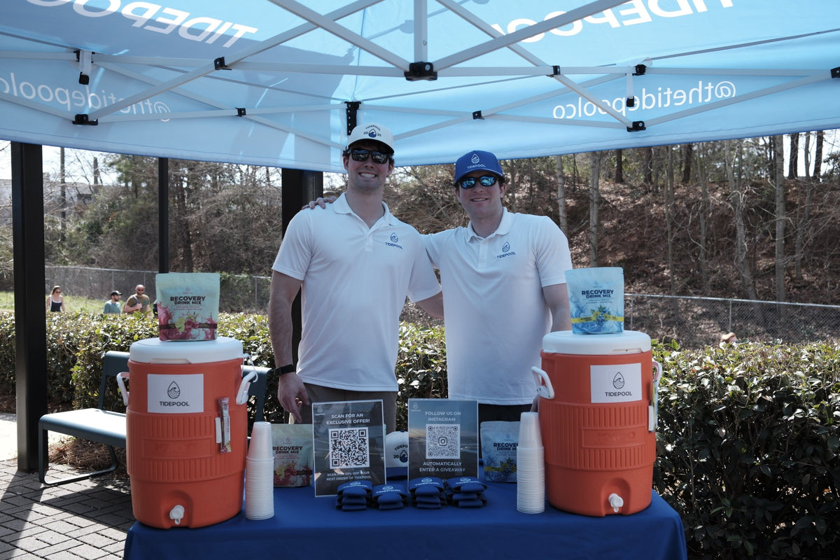 Two men standing behind a table with orange coolers and branded items under a blue canopy.